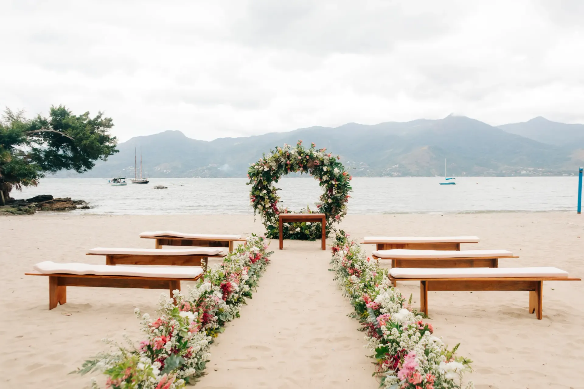 Noiva ao lado do arco de flores em cerimônia à beira-mar, com barcos, montanhas e mar azul ao fundo