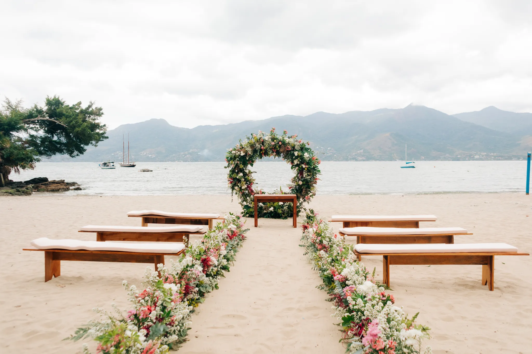 Noiva ao lado do arco de flores em cerimônia à beira-mar, com barcos, montanhas e mar azul ao fundo